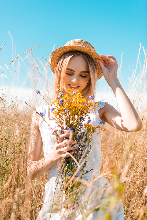 selective focus of blonde woman in white dress touching straw hat while holding wildflowers in grassy field