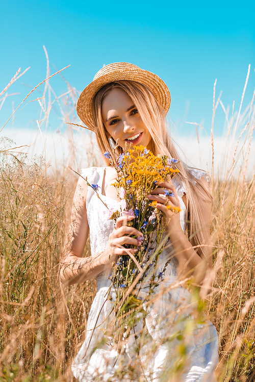 selective focus of sensual woman in white dress and straw hat  while holding wildflowers in meadow