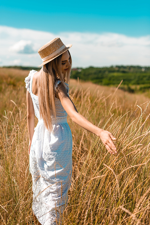 young blonde woman in white dress and straw hat touching spikelets while standing in field