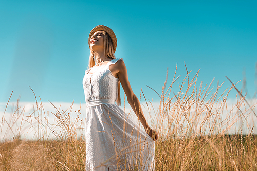 selective focus of young woman in straw hat touching white dress while standing with closed eyes in grassy meadow