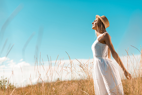 selective focus of stylish woman in white dress and straw hat standing with outstretched hands and closed eyes in grassland