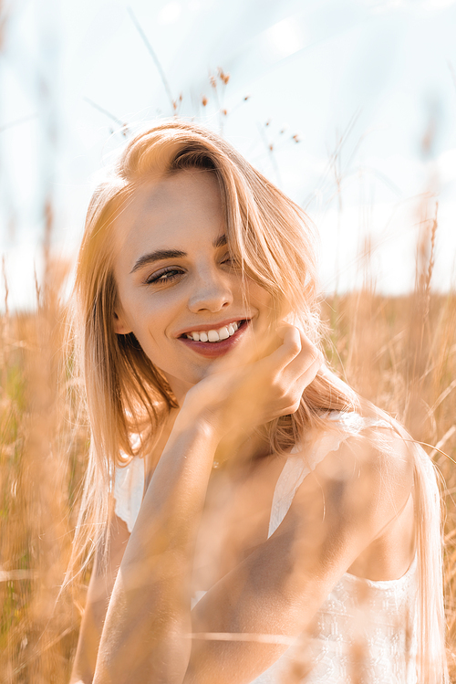 selective focus of young blonde woman holding hand near face while looking away in grassland