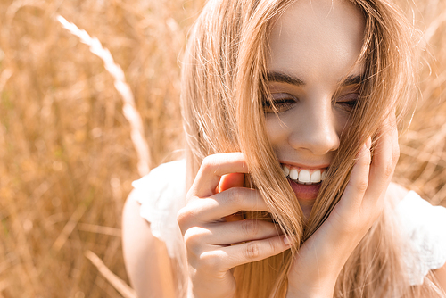 excited, young blonde woman touching hair near grass in meadow
