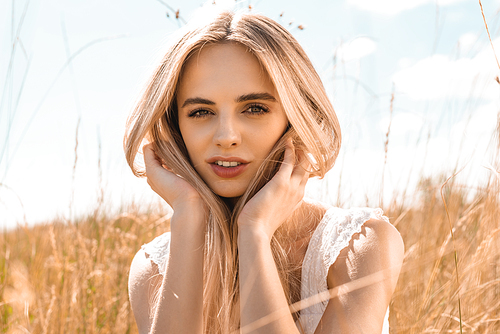 selective focus of dreamy blonde woman touching hair and  against blue sky in meadow