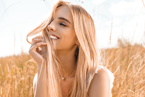 sensual blonde woman touching hair and looking away near grass in field