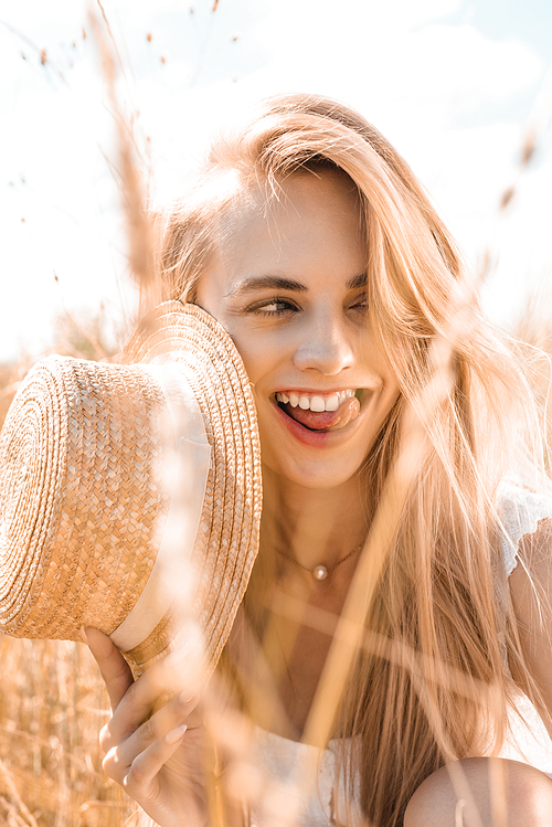 selective focus of blonde woman sticking out tongue while holding straw hat and looking away in field