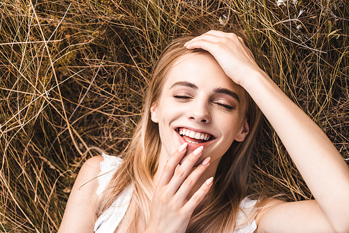 top view of excited blonde woman touching lips and forehead while lying on grass with closed eyes