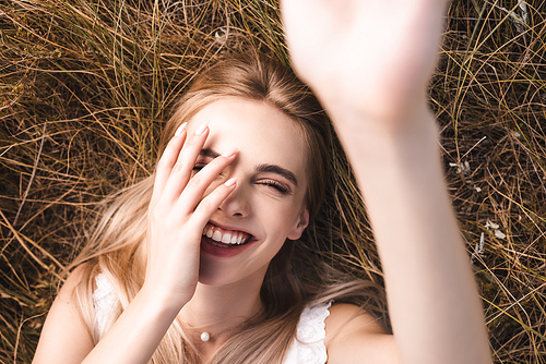 selective focus of excited blonde woman touching face and  while lying on grass with raised hand, top view