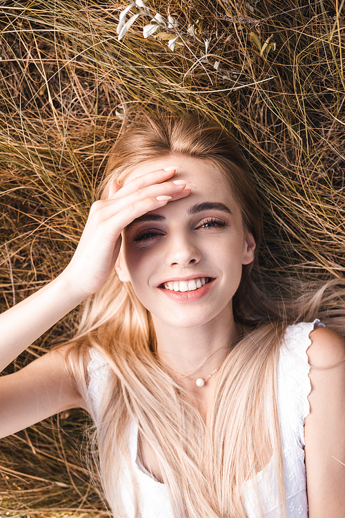 top view of excited blonde woman touching forehead and  while lying on green grass