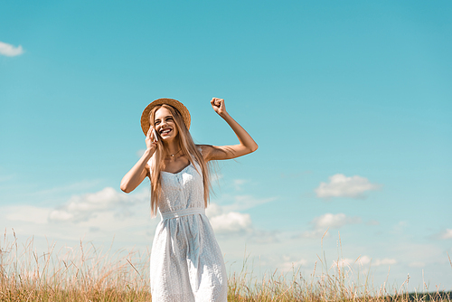 excited blonde woman in white dress and straw hat showing winner gesture while talking on smartphone against blue sky