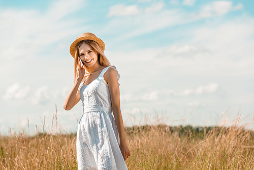 young woman in white dress and straw hat  while talking on smartphone in meadow against blue sky