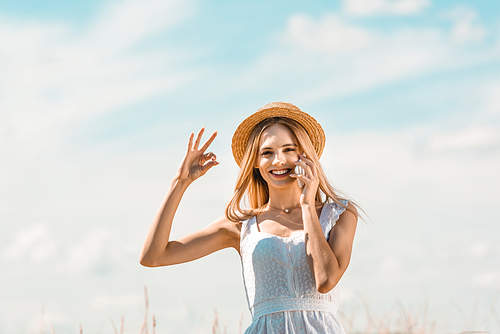 young, excited woman in white dress and straw hat showing okay gesture while talking on smartphone against blue sky