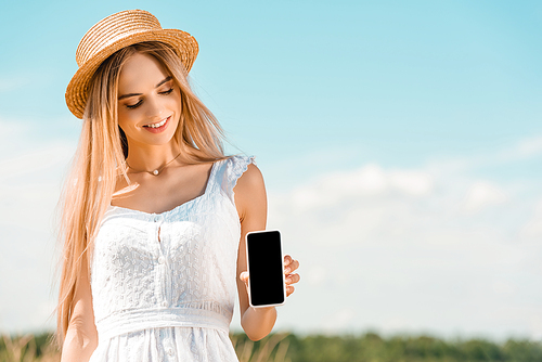blonde woman in white dress and straw hat showing smartphone with blank screen against blue sky