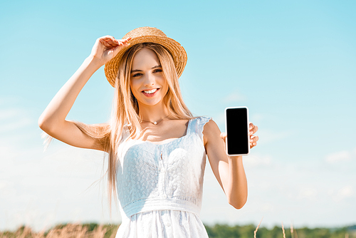 young blonde woman in white dress touching straw hat while showing smartphone with blank screen against blue sky