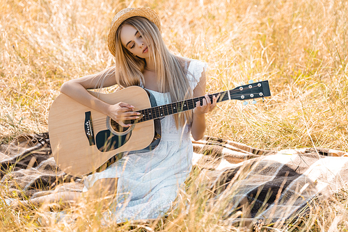 dreamy woman in white dress and straw hat playing acoustic guitar while sitting on blanket in field