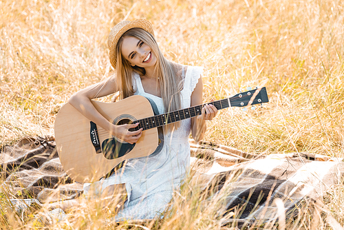 selective focus of young woman in white dress and straw hat  while playing acoustic guitar on blanket in field