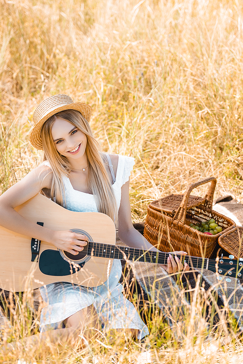 selective focus of blonde woman in straw hat  while playing acoustic guitar on blanket in meadow, high angle view
