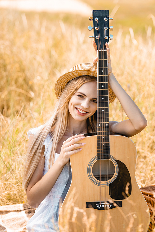 selective focus of young blonde woman  while sitting with acoustic guitar in grassy field