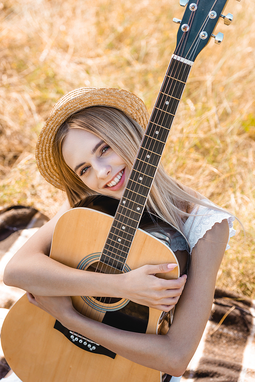 high angle view of blonde woman in straw hat sitting on blanket in field with acoustic guitar
