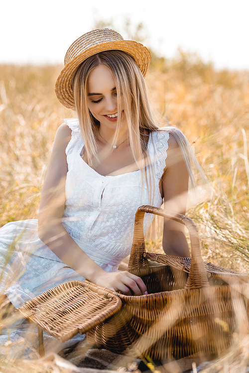 blonde woman in white dress and straw hat sitting near wicker basket in field