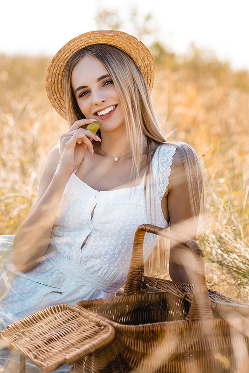 selective focus of blonde woman in white dress and straw hat holding ripe grape while  in field
