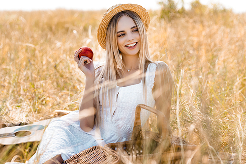 selective focus of woman in white dress and straw hat holding ripe apple while sitting in field