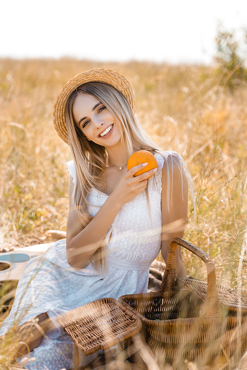 selective focus of stylish woman in white dress and straw hat holding orange while sitting in field near wicker basket