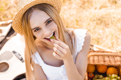 high angle view of blonde woman in straw hat eating ripe grape and  on picnic in field