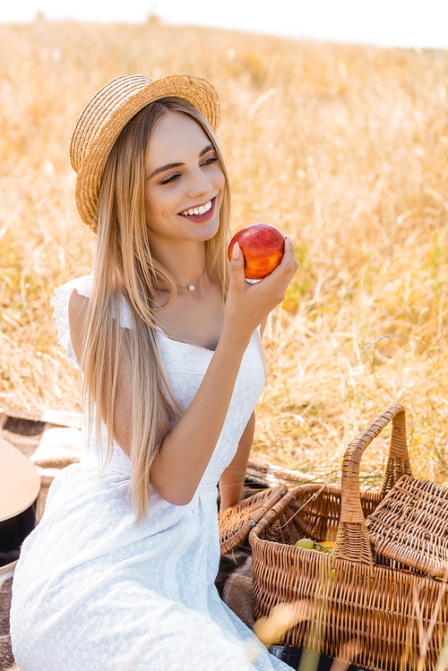 selective focus of blonde woman in straw hat and white dress sitting on blanket near wicker basket and holding ripe apple