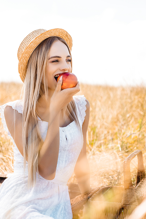 selective focus of blonde woman in straw hat eating ripe apple and looking away in field