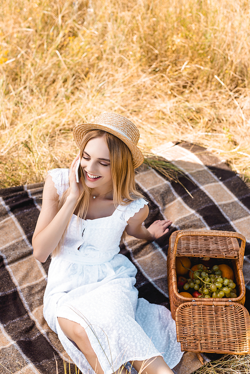 high angle view of blonde woman in summer outfit talking on smartphone near wicker basket with fruits