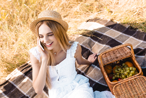 high angle view of blonde woman in white dress and straw hat talking on smartphone near wicker basket with fruits