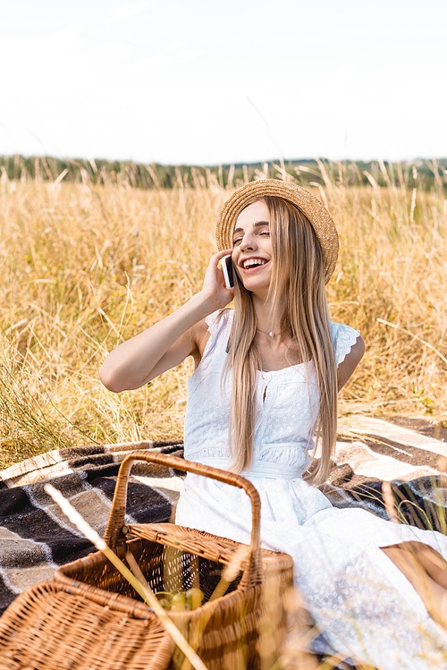 selective focus of excited woman in white dress and straw hat talking on smartphone near wicker
