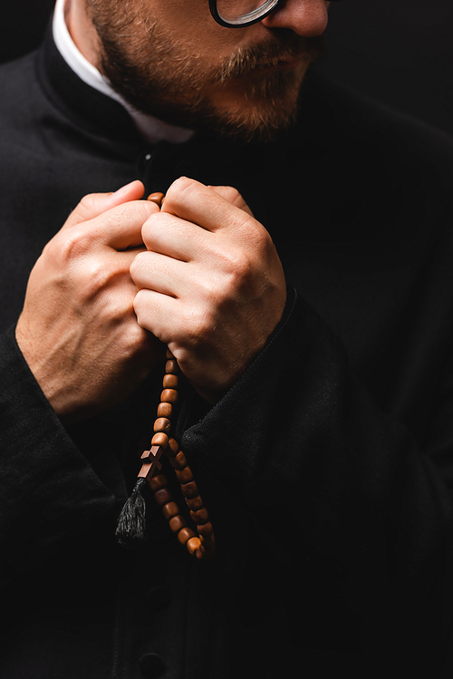 cropped view of bearded priest holding rosary beads in hands and praying isolated on black
