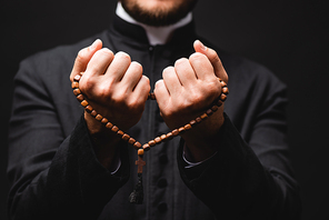 selective focus of pastor holding rosary beads in hands and praying isolated on black