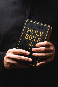 partial view of priest holding holy bible in hands isolated on black