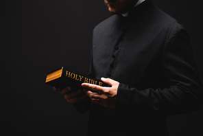 cropped view of bearded priest holding holy bible in hands isolated on black