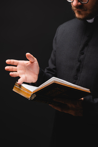 cropped view of priest holding holy bible and gesturing isolated on black