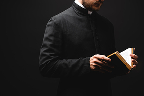 partial view of bearded priest holding holy bible in hands isolated on black
