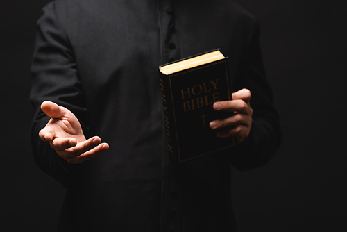partial view of priest holding holy bible while gesturing isolated on black