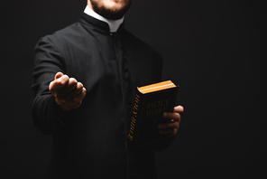 cropped view of priest holding holy bible while showing fist isolated on black