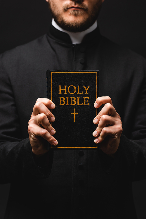cropped view of priest holding book with holy bible lettering isolated on black