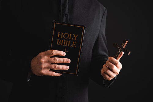 cropped view of priest holding cross and holy bible isolated on black