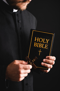 cropped view of bearded priest holding cross and holy bible isolated on black