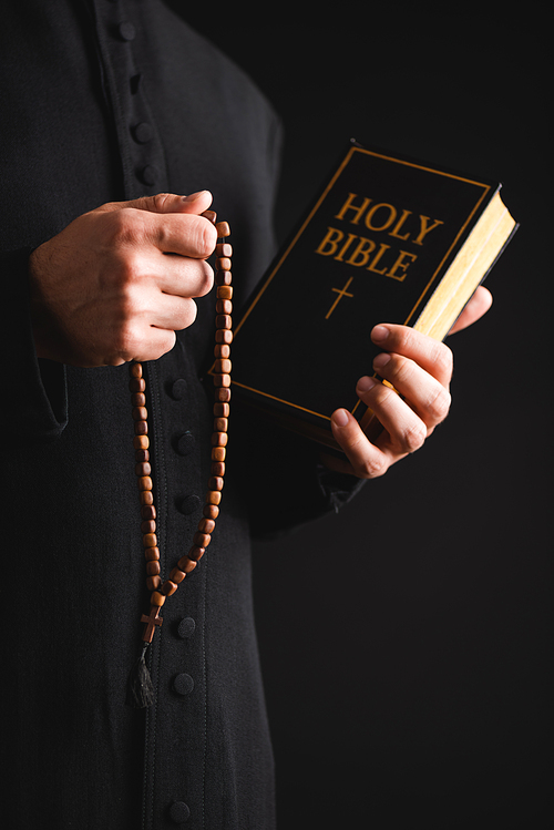cropped view of priest holding holy bible and rosary beads in hands isolated on black