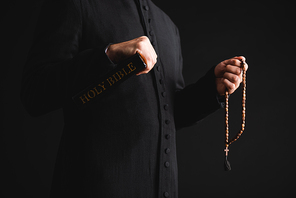 cropped view of pastor holding holy bible and rosary beads in hands isolated on black