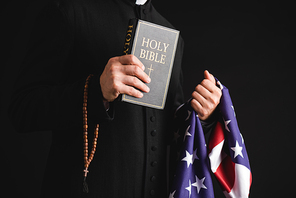 cropped view of priest holding holy bible, american flag and rosary beads isolated on black