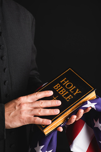 cropped view of priest holding holy bible and american flag isolated on black