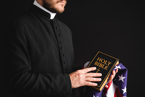 partial view of priest holding holy bible and american flag isolated on black