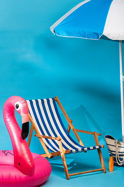 striped deck chair near inflatable flamingo, sunscreen, beach bag and umbrella on blue background